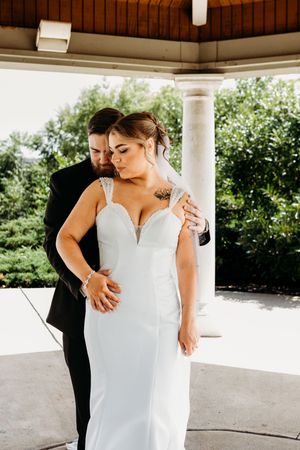 Groom in black suit embraces bride in fitted white gown under a wooden gazebo outdoors.