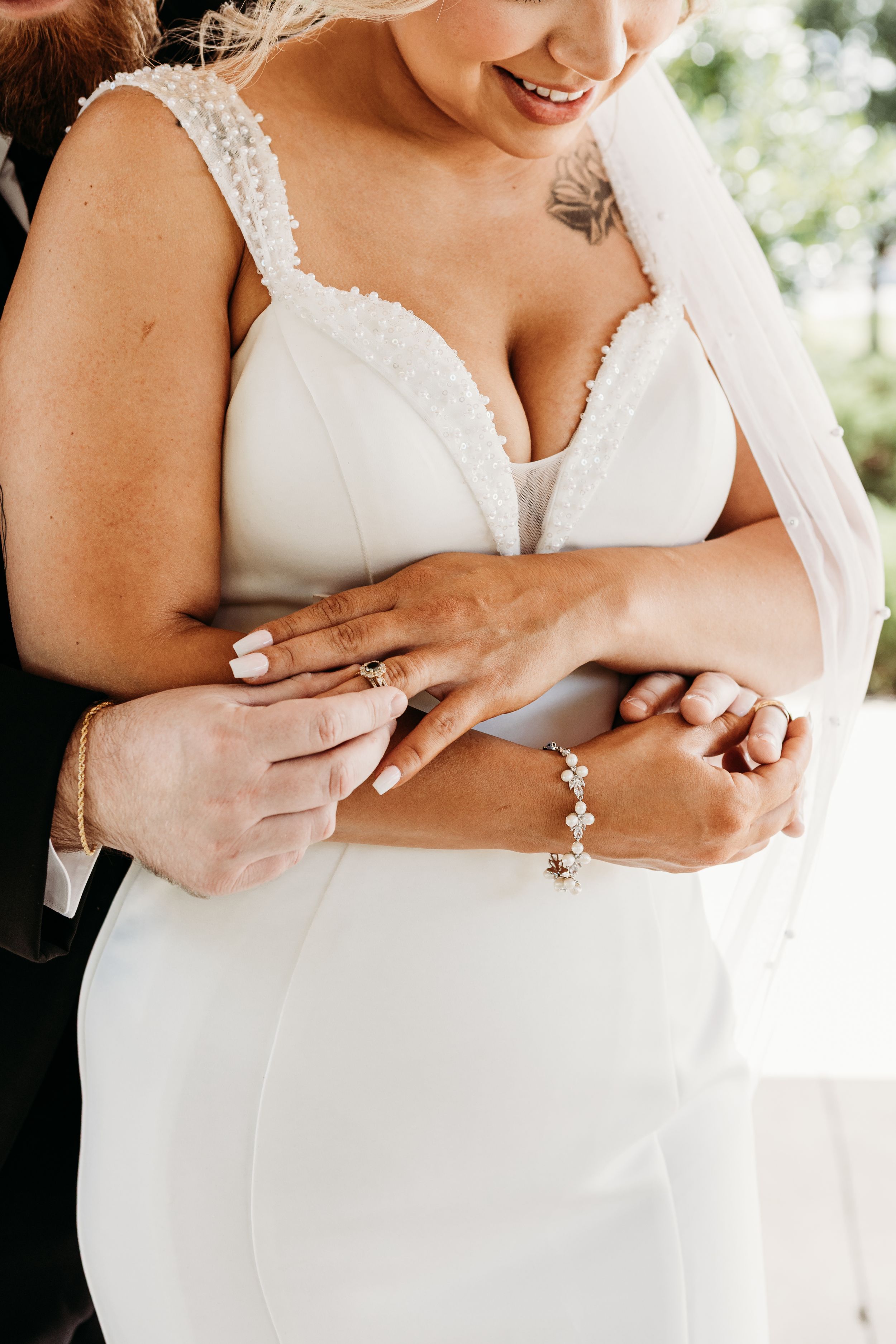 Close-up of groom's arms wrapped around bride in white dress, highlighting wedding jewelry.