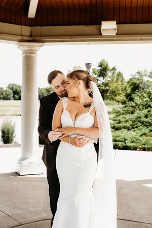 Groom tenderly kisses bride's cheek while holding her close under an elegant outdoor pavilion.