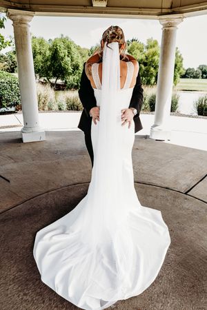 Bride in a stunning white mermaid gown with veil embraced by groom under a pavilion.