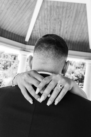 Black and white close-up of bride's hands with wedding ring resting on groom's shoulders.