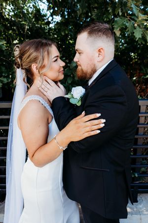 Bearded groom gently cups brides face as they gaze at each other near a garden fence.