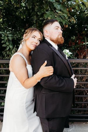 Bride in white gown leans on grooms back as they pose together in a lush garden.
