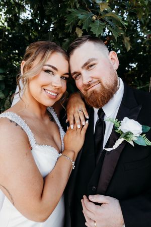 Smiling bride and bearded groom pose cheek-to-cheek showing wedding rings in lush garden setting.