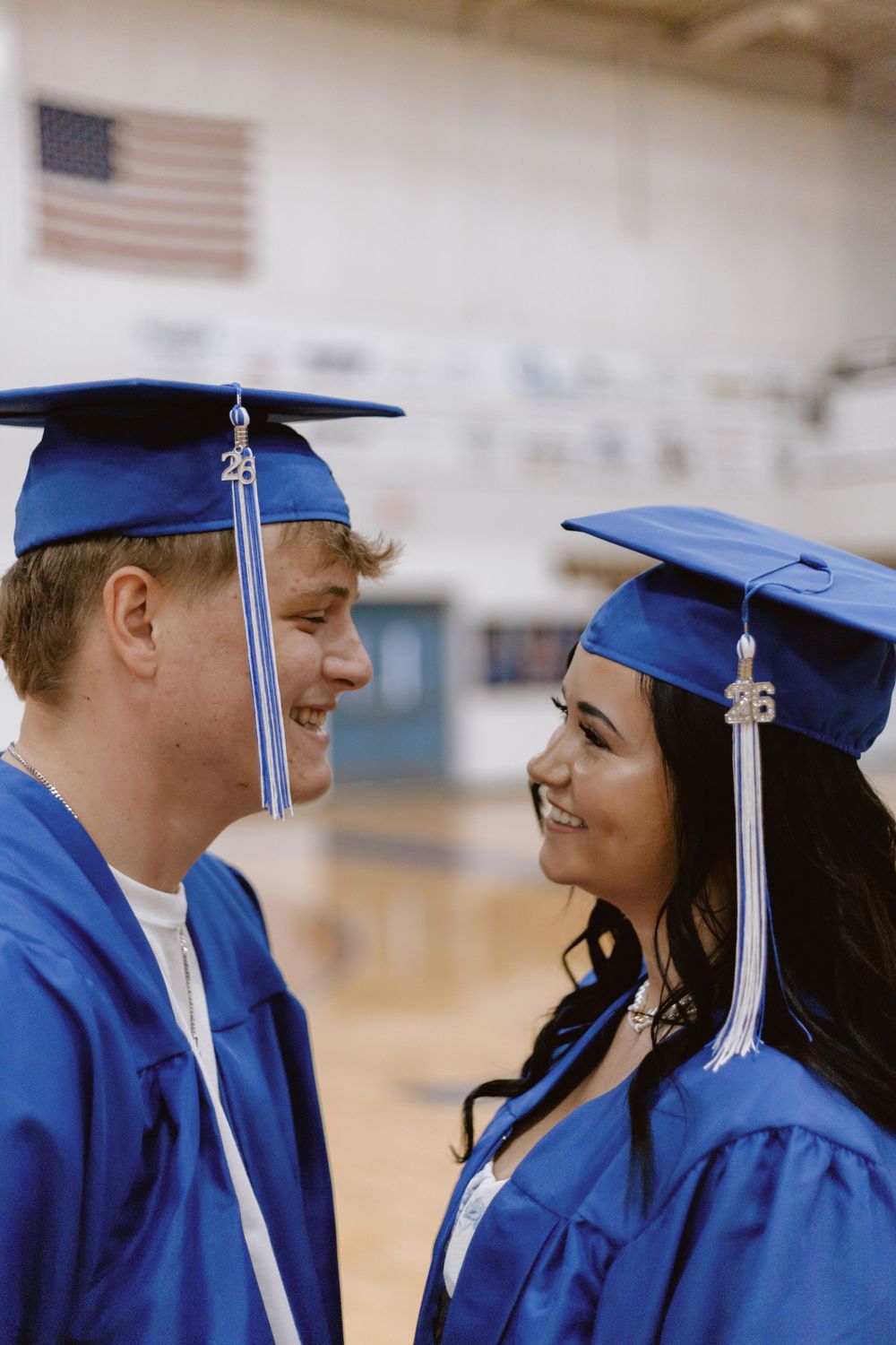 Two high school graduates in blue caps and gowns smiling at each other in a gymnasium.