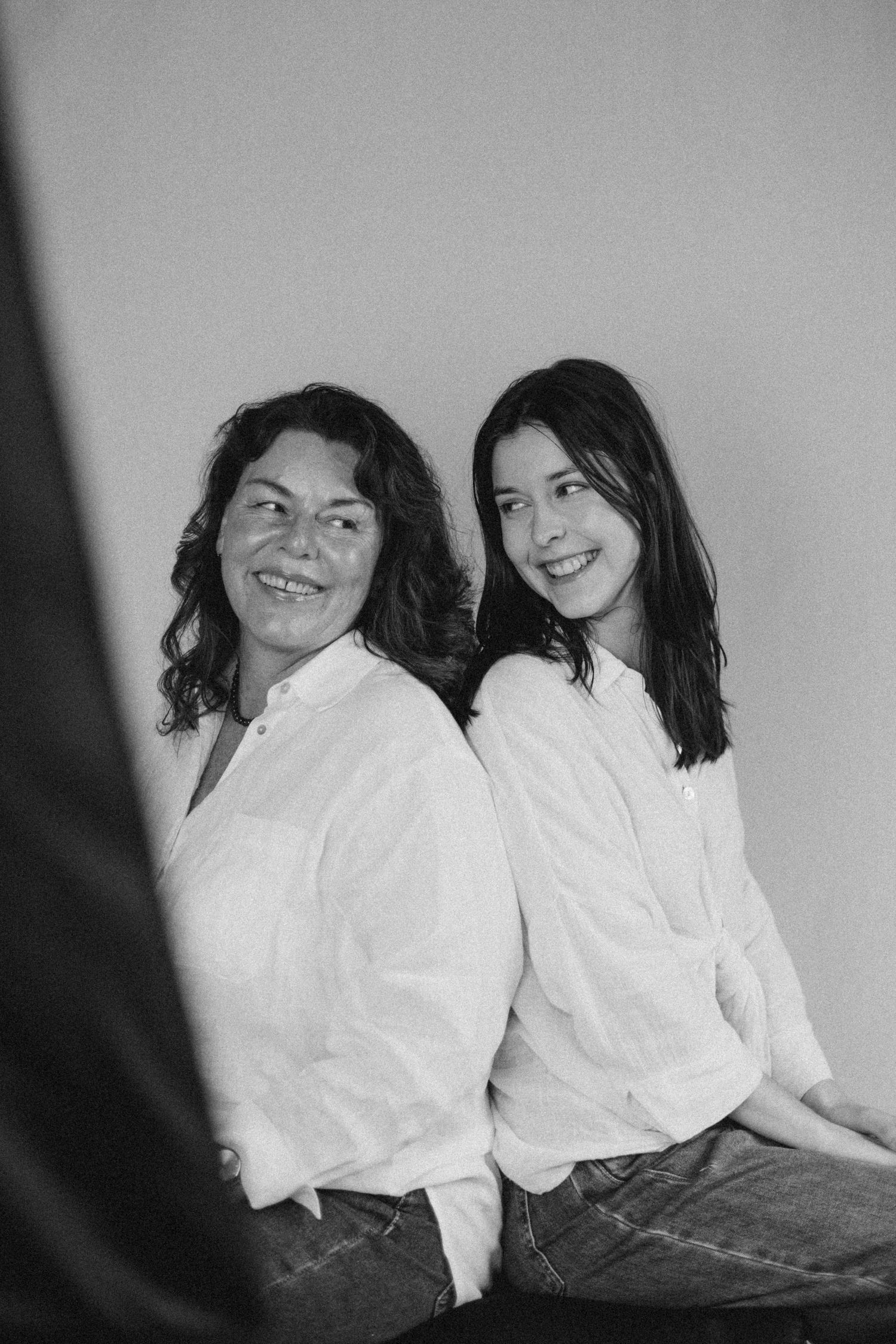 Two women in white shirts sitting back-to-back, smiling, in a black and white portrait photo.