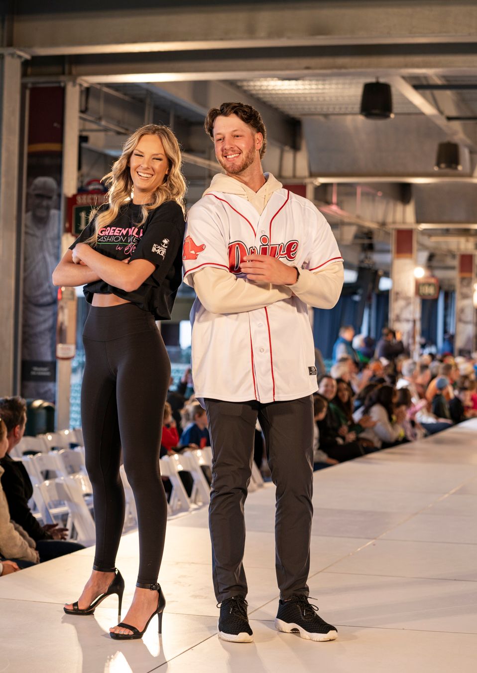 Two people pose on a fashion runway, one wearing a black athletic outfit and one in a baseball jersey and jeans.