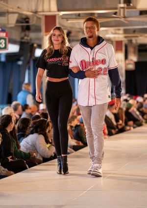 People walking on a fashion runway wearing casual sportswear and baseball-themed outfits in an indoor venue.