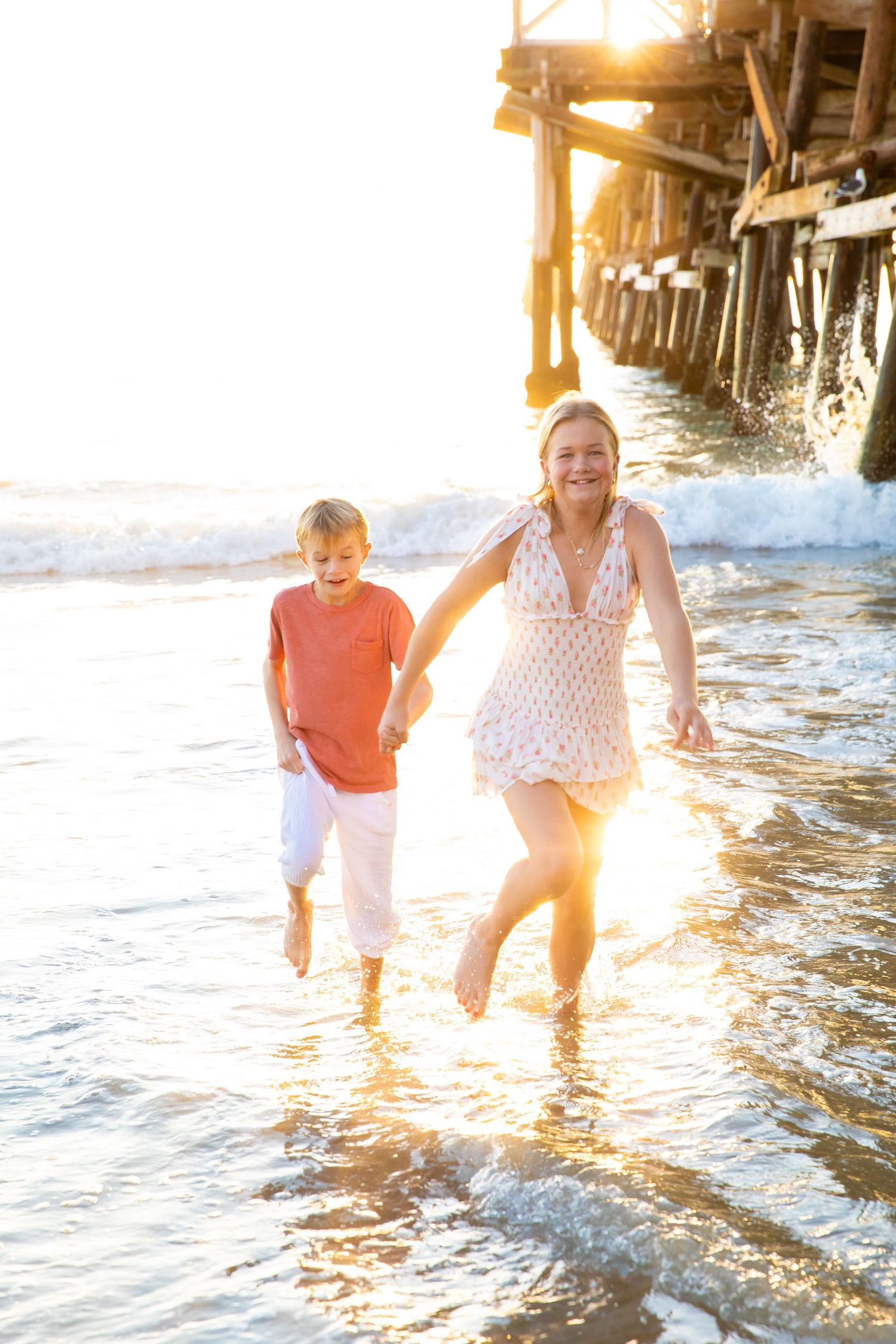 Two people splash and play in shallow ocean waves during golden sunset under a pier.