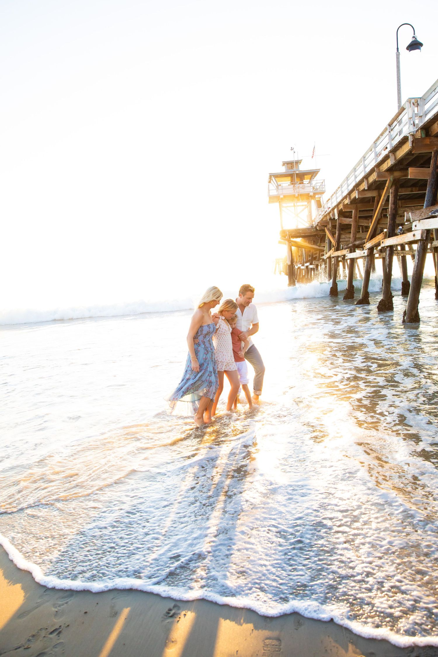 A couple plays in the waves at sunset near a pier, creating a romantic silhouette against the golden light.