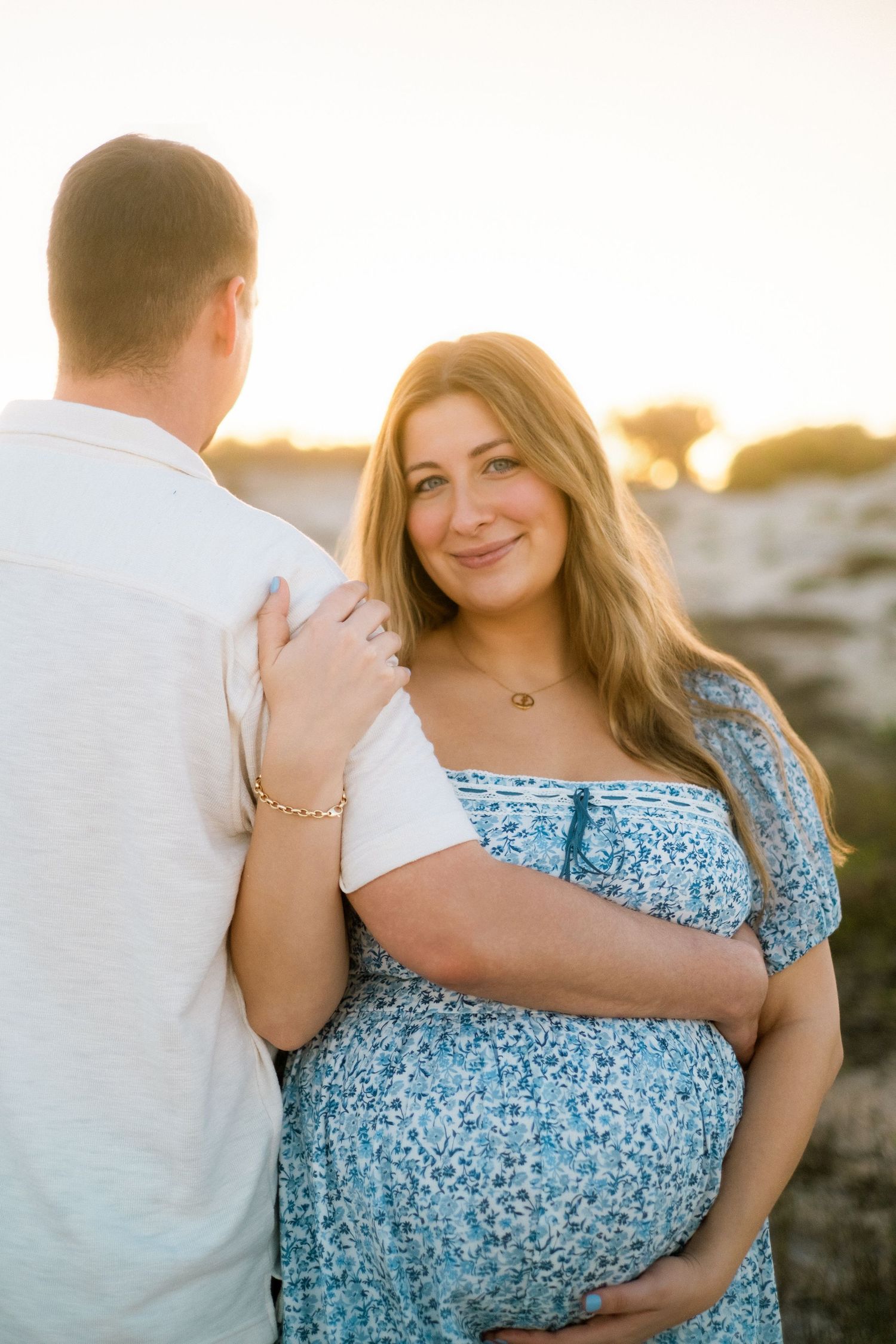 Smiling pregnant woman in blue floral dress embracing her partner during golden hour outdoors.
