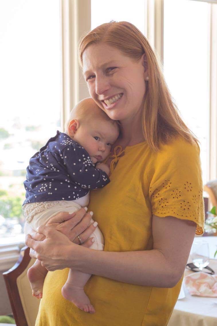 A baby wearing a yellow knit hat and white outfit smiles at the camera.