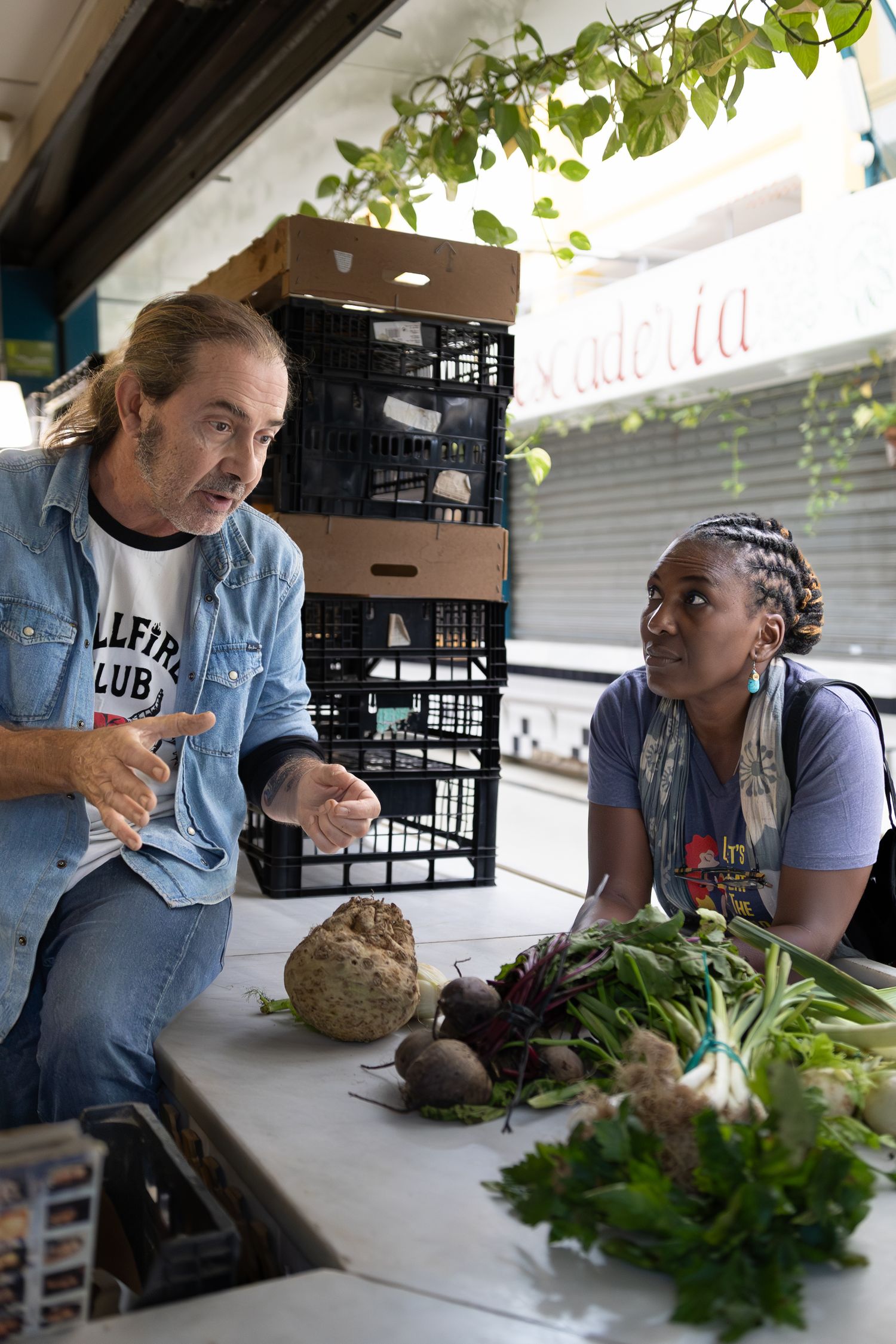 Two people discuss produce and supplies at a market stand with fresh vegetables and storage crates on display.