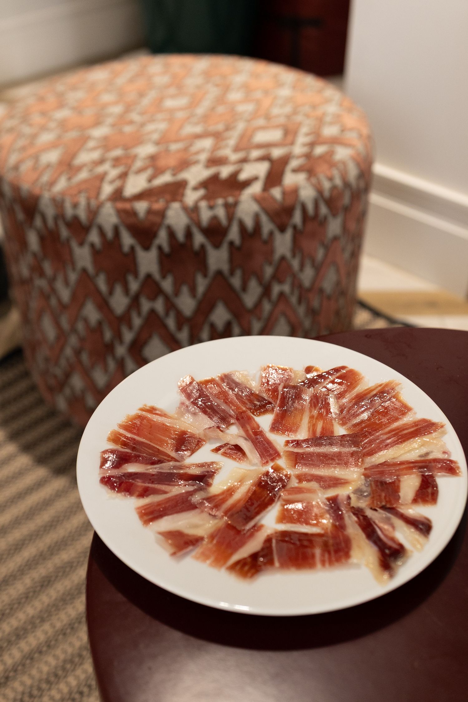 A plate of thinly sliced Spanish jamon iberico ham arranged in a circular pattern with a patterned ottoman in background.