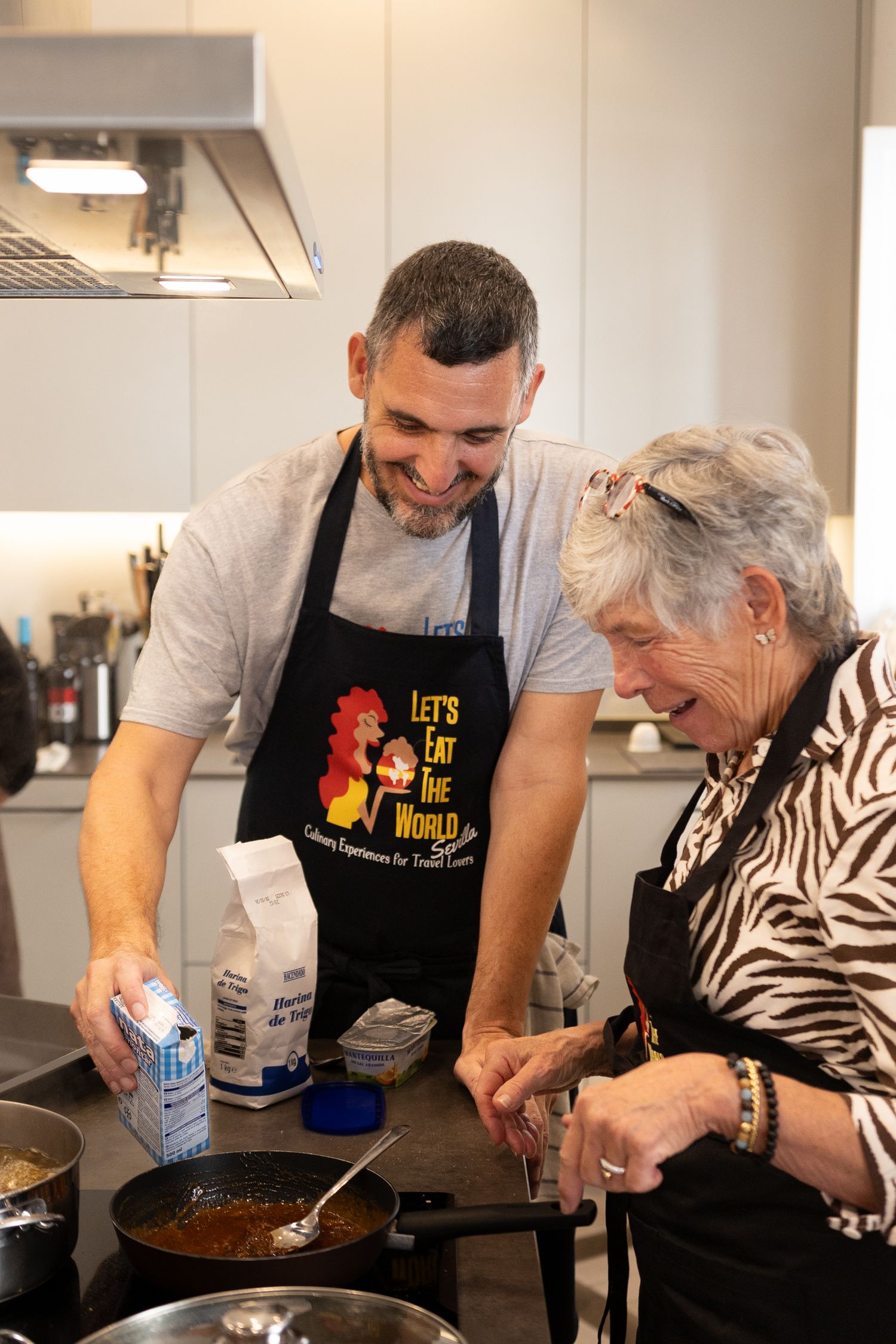 A cooking instructor demonstrates techniques to an elderly student in a kitchen setting.