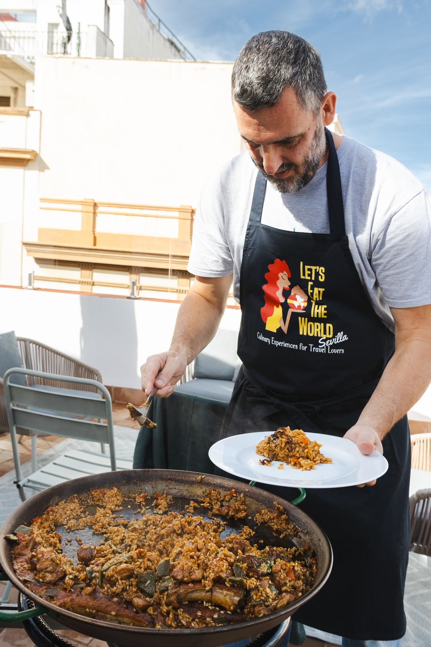 A chef in a black apron prepares paella in a large traditional pan on an outdoor terrace.