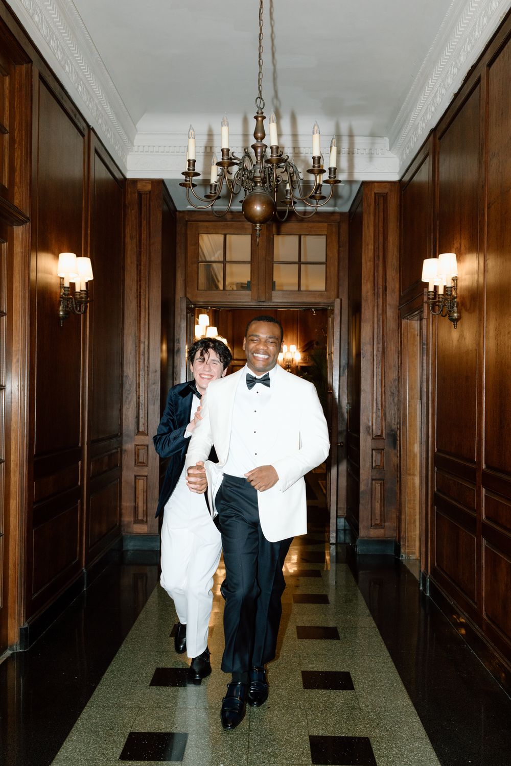 People in formal attire walk through an elegant wood-paneled hallway with chandeliers and wall sconces.