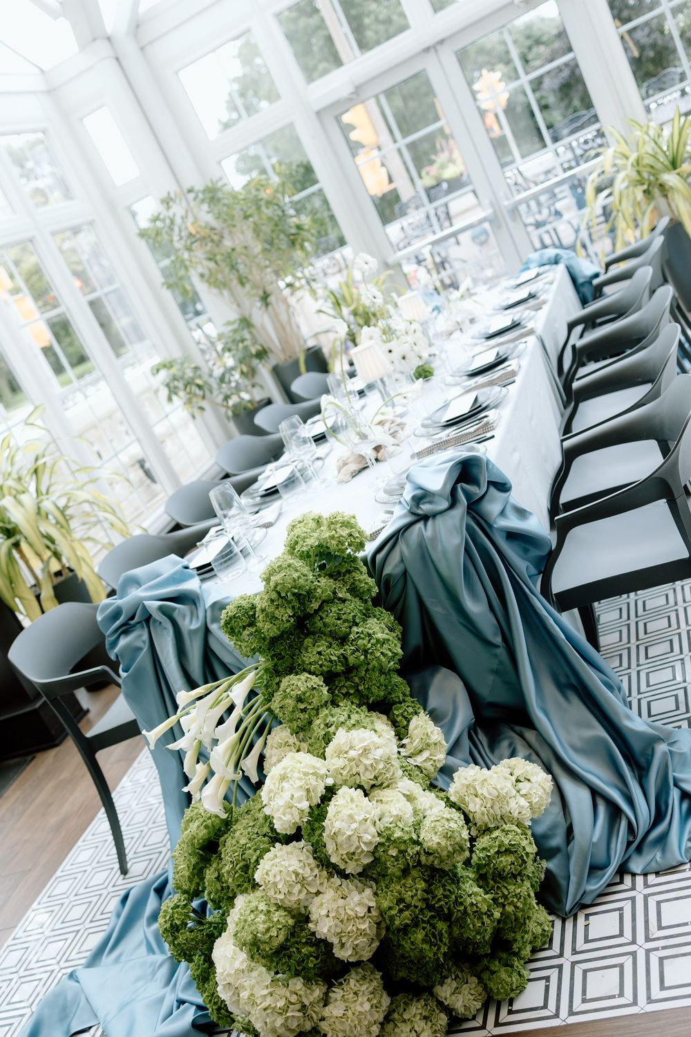 Elegant wedding table setting with teal linens and white hydrangea centerpieces in a bright conservatory space.