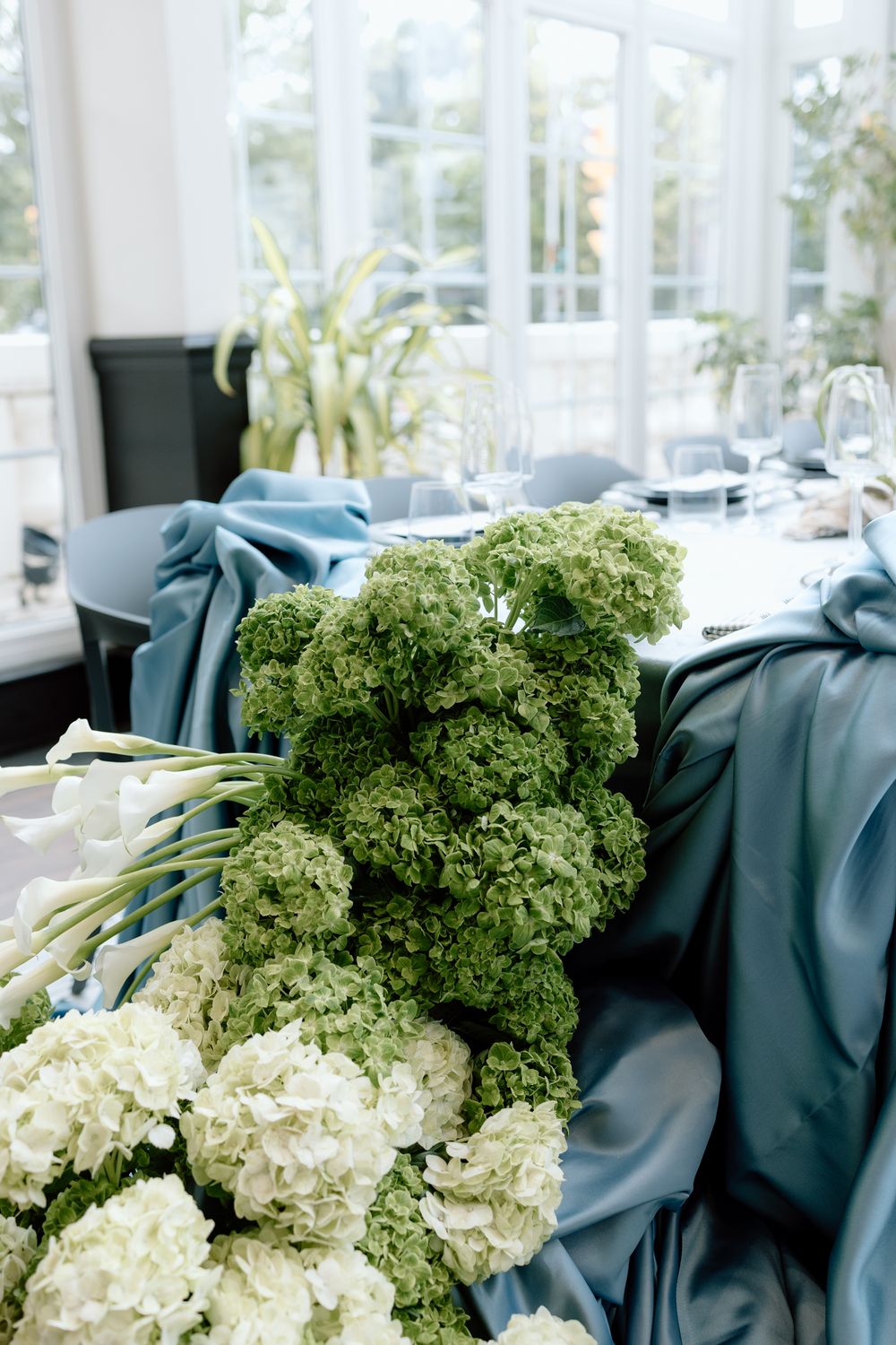Green hydrangeas and white floral arrangements rest against teal blue fabric in an elegant event setting.