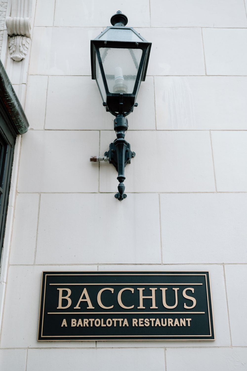 Exterior signage for Bacchus restaurant with black and gold lettering mounted on a white building wall with lanterns.