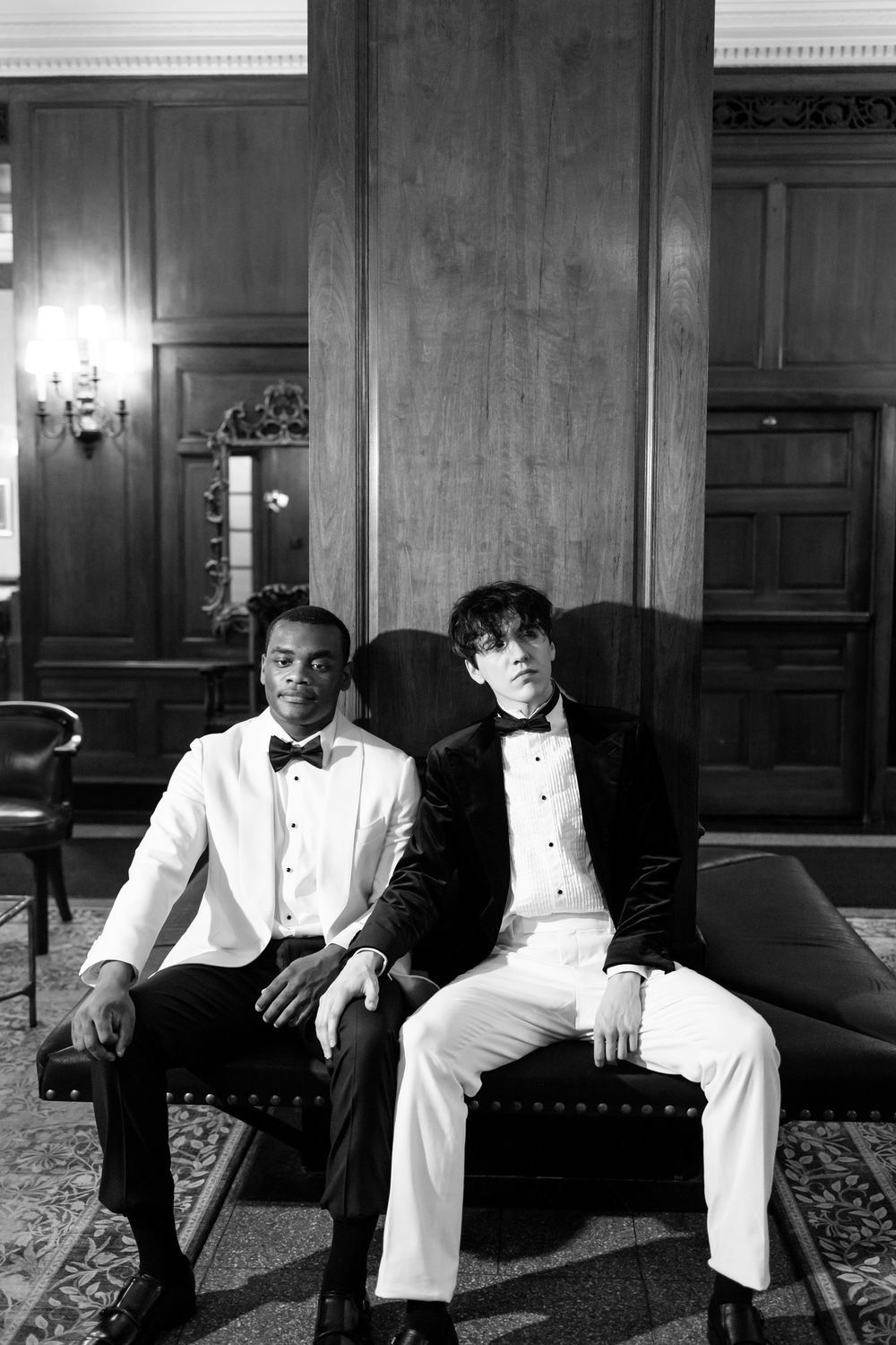 A black and white photograph shows men in formal white tuxedos seated on an elegant couch in an ornate room.