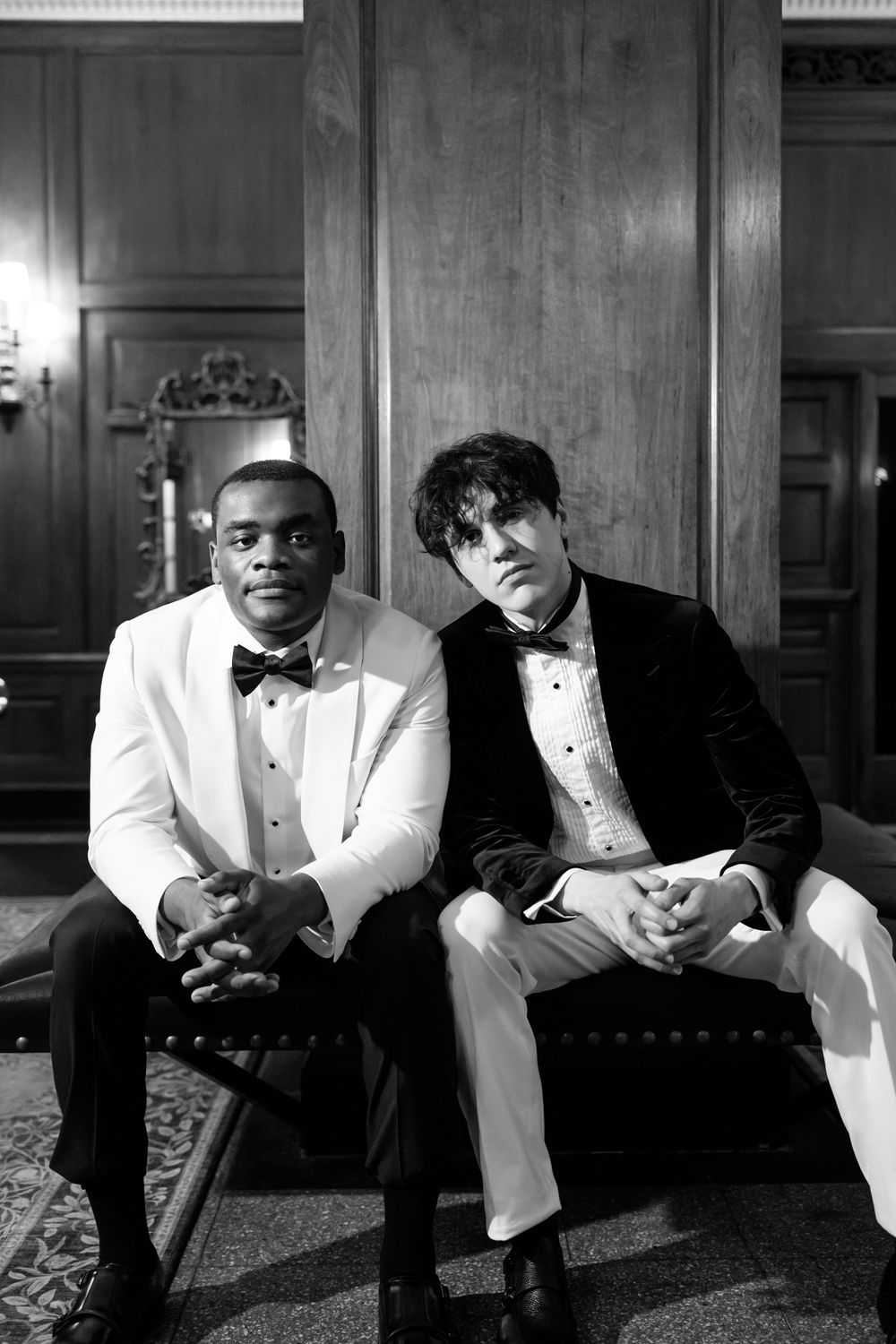Two people in formal tuxedos sit together on an elegant bench in a dimly lit room.