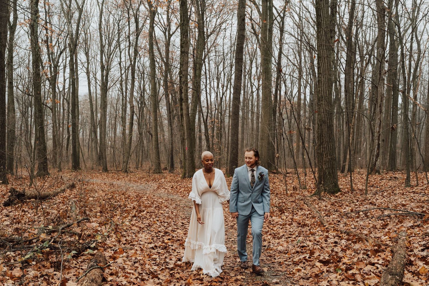 A couple walks hand in hand through a forest filled with bare trees and fallen autumn leaves.