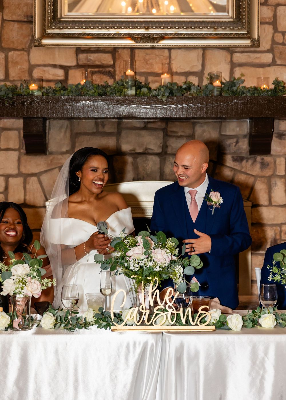 Head table at wedding reception with white linens and floral arrangements against stone wall.