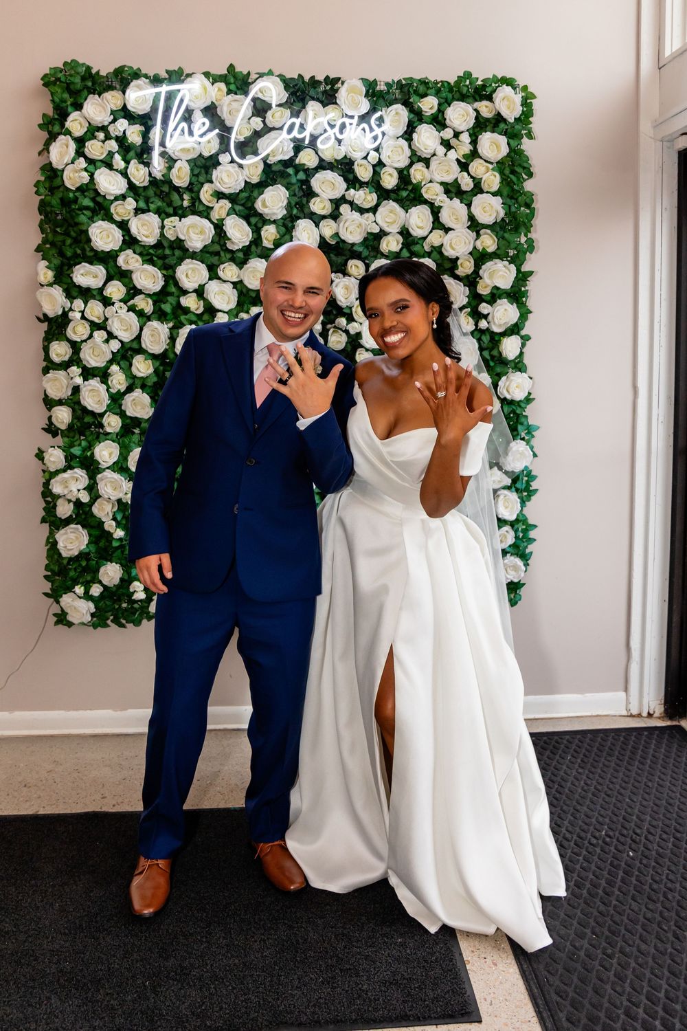 Wedding couple poses playfully in front of white rose wall backdrop.