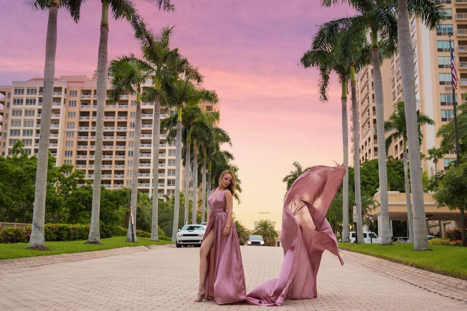 Two figures in flowing pink dresses pose on a palm-lined street at sunset with high-rise buildings in the background.