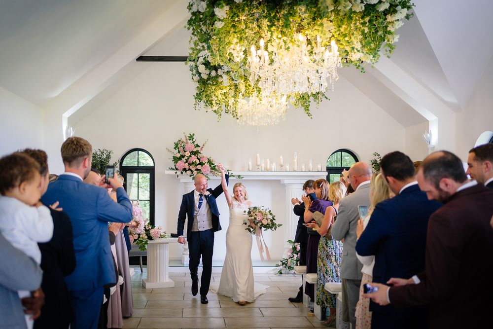 Wedding ceremony in progress under illuminated floral chandelier in bright chapel setting. Woodhill Hall.