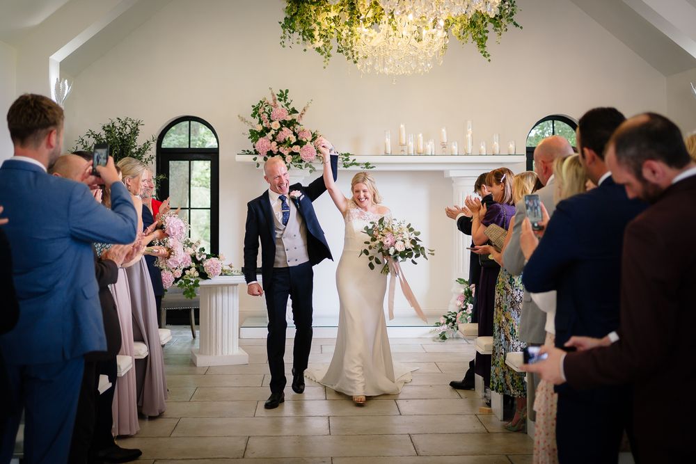 Wedding guests celebrate during an elegant indoor reception with a chandelier and natural light. Woodhill Hall.