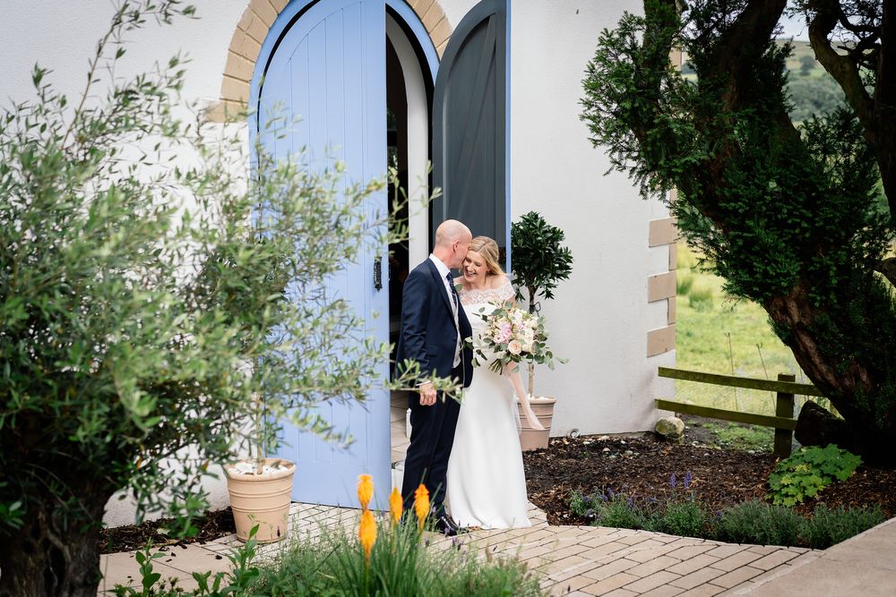 Outdoor wedding ceremony setup with white chairs and floral arrangements against a stone building. Woodhill Hall.