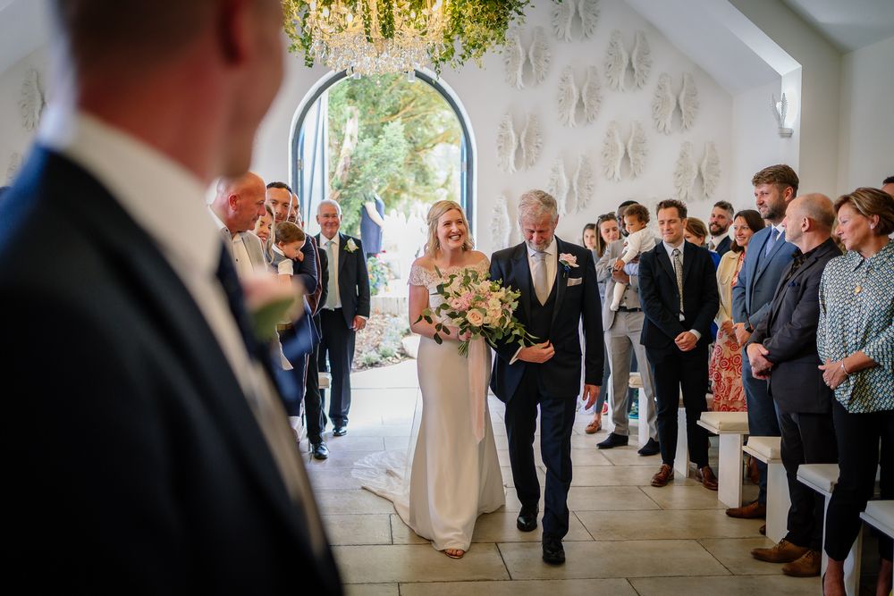 Wedding ceremony guests line up along white walls of chapel interior. Woodhill Hall.