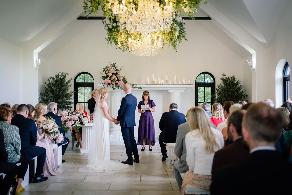Wedding ceremony in progress inside bright white chapel with hanging floral arrangements. Woodhill Hall.