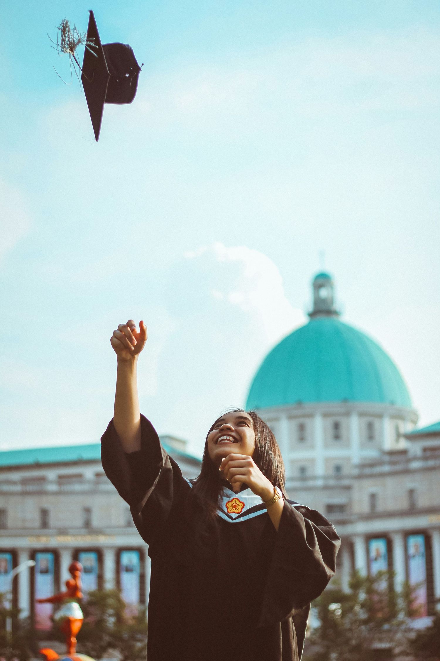 Graduates in black caps and gowns celebrate by tossing their mortarboards into the air against a blue sky and domed building.