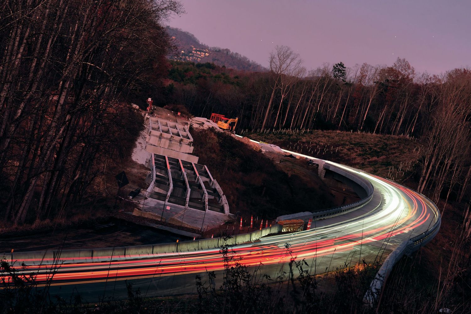 Long exposure photograph of light trails on a winding mountain road at dusk with a concrete dam structure visible.