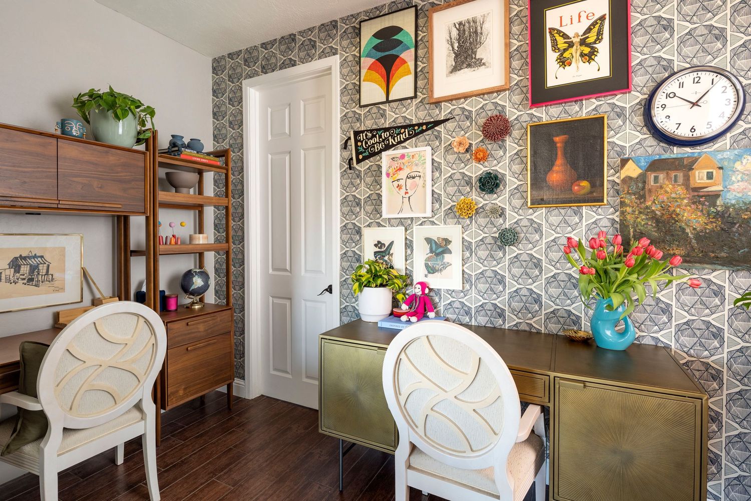 Eclectic home office with patterned wallpaper, white chairs, wooden shelving, and colorful framed botanical artwork on the wall.