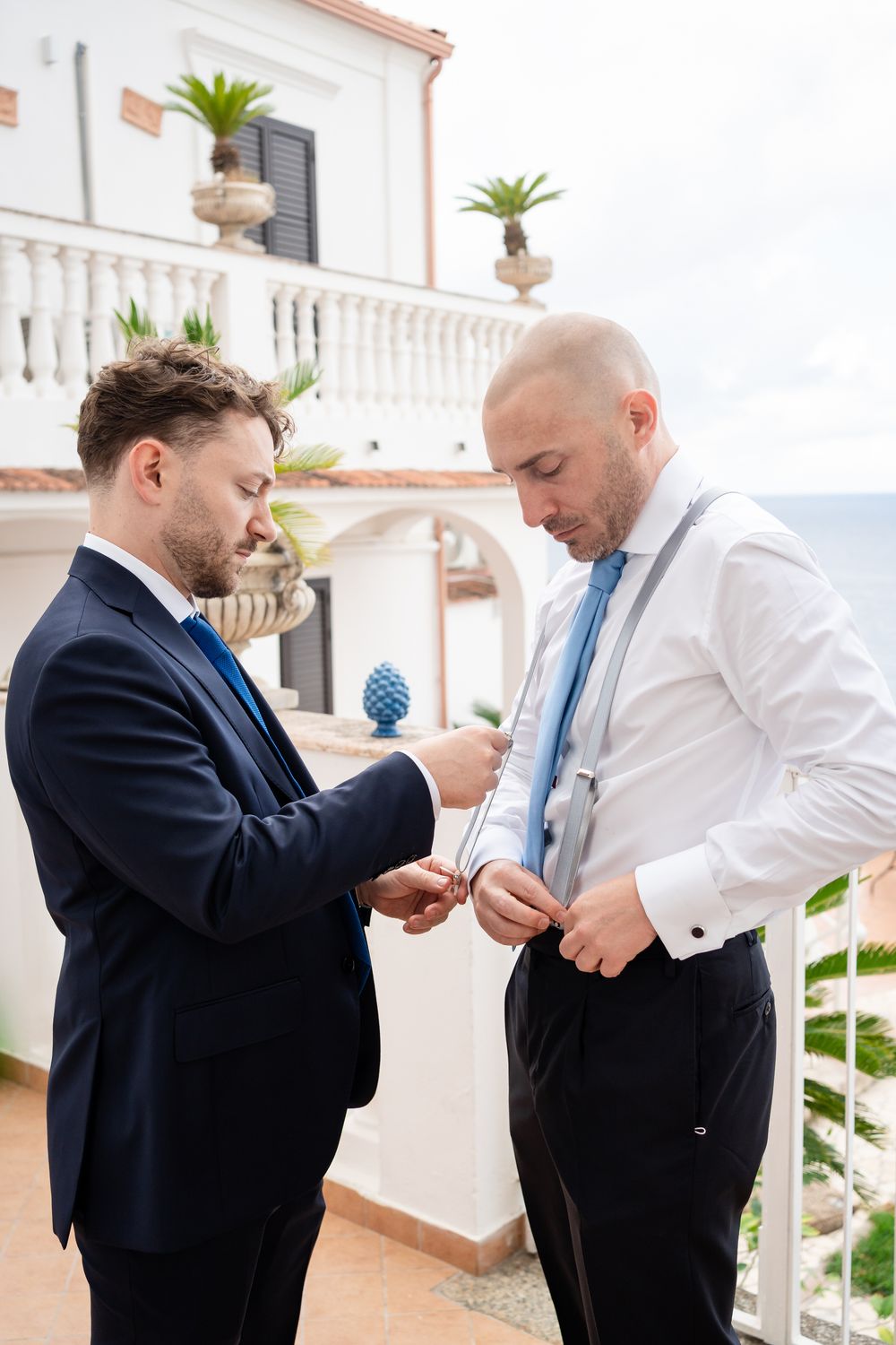 Two men in formal attire help each other get dressed on an elegant terrace with potted palms and ocean views.