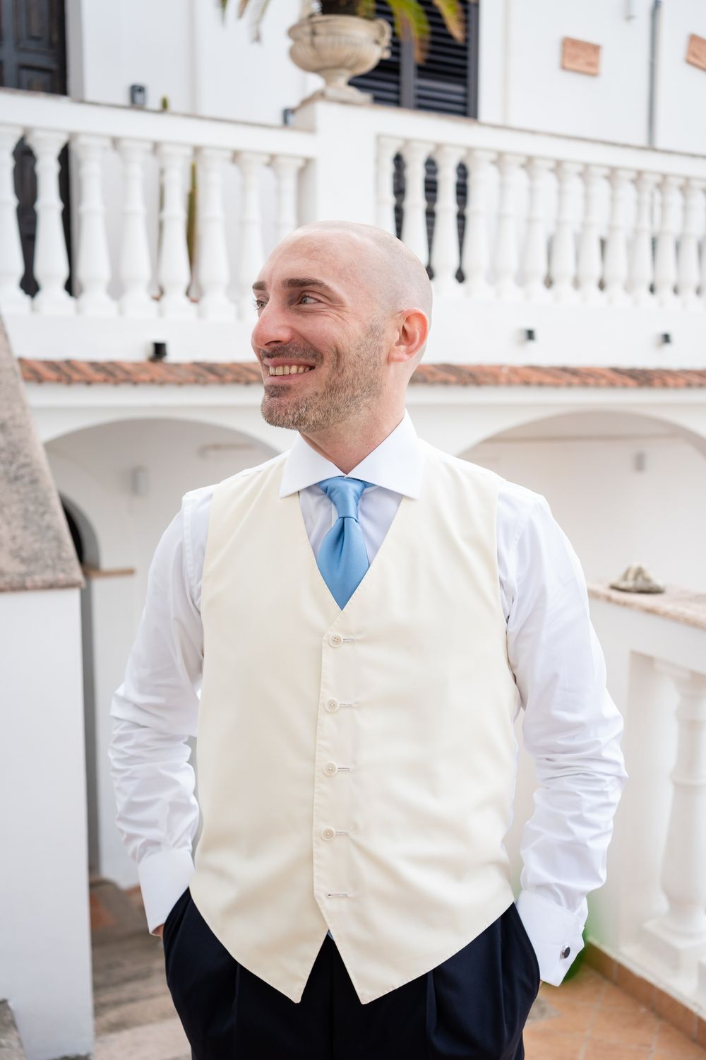 A groom in a cream vest and blue tie smiles joyfully while standing on a white balcony.