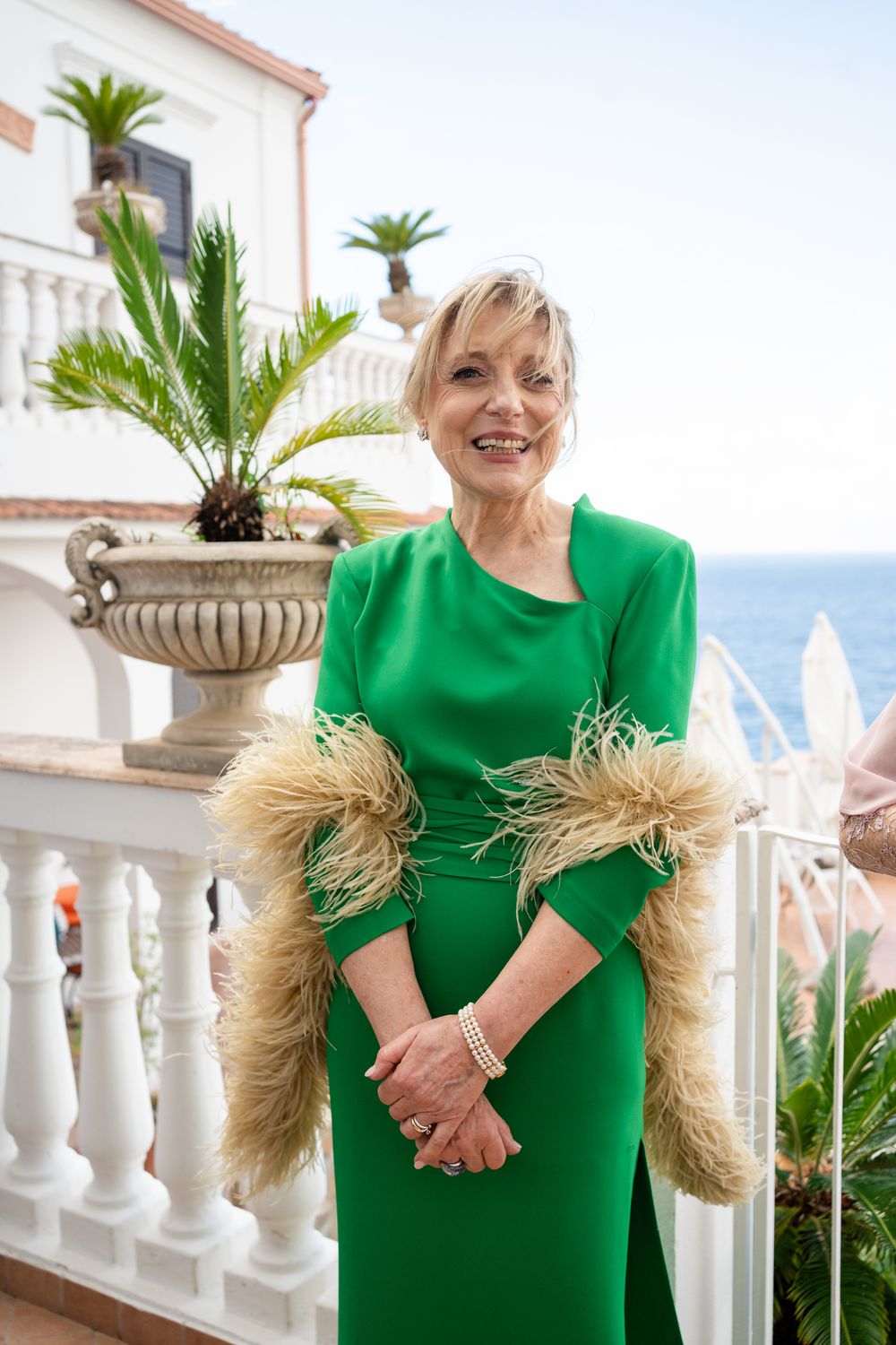 Person in bright green dress with straw-like sleeves poses on Mediterranean balcony with white columns and palm trees.