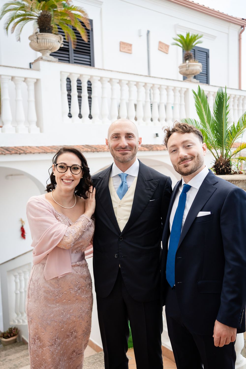 Group photo of wedding party in formal attire on elegant balcony with white columns and palm trees.