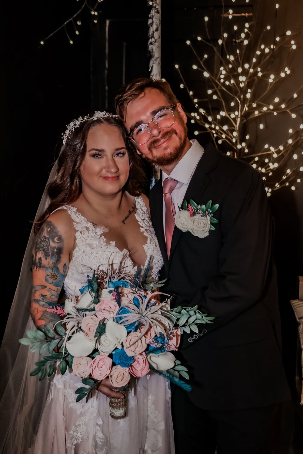 A wedding couple poses together with a beautiful floral bouquet under twinkling lights.