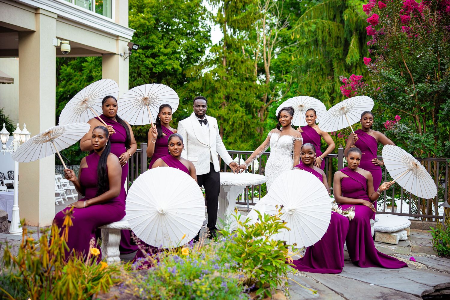 Wedding party in magenta dresses poses with white parasols in a garden setting during an outdoor celebration.