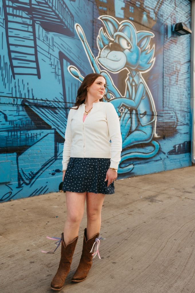 A casual outfit featuring brown cowboy boots and a white sweater is styled against a blue graffiti wall backdrop.