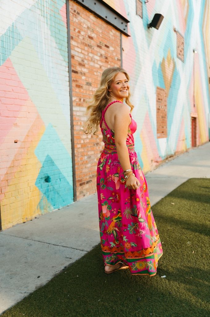 Person in vibrant pink floral dress poses playfully against colorful geometric wall mural in urban setting.