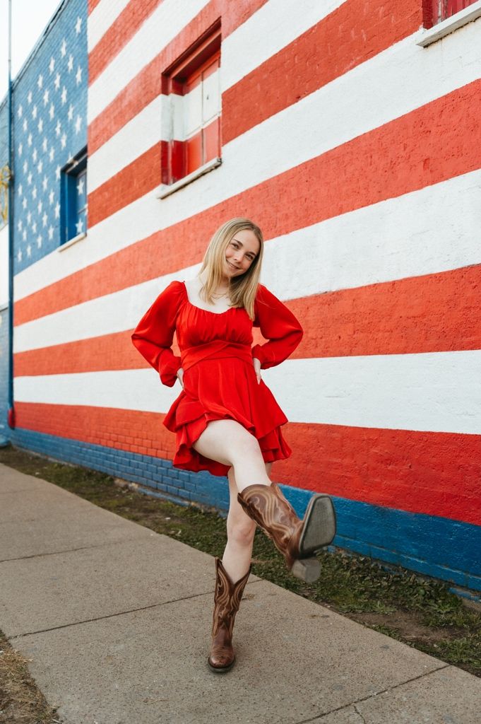Woman in red dress poses against American flag mural wall in brown cowboy boots.