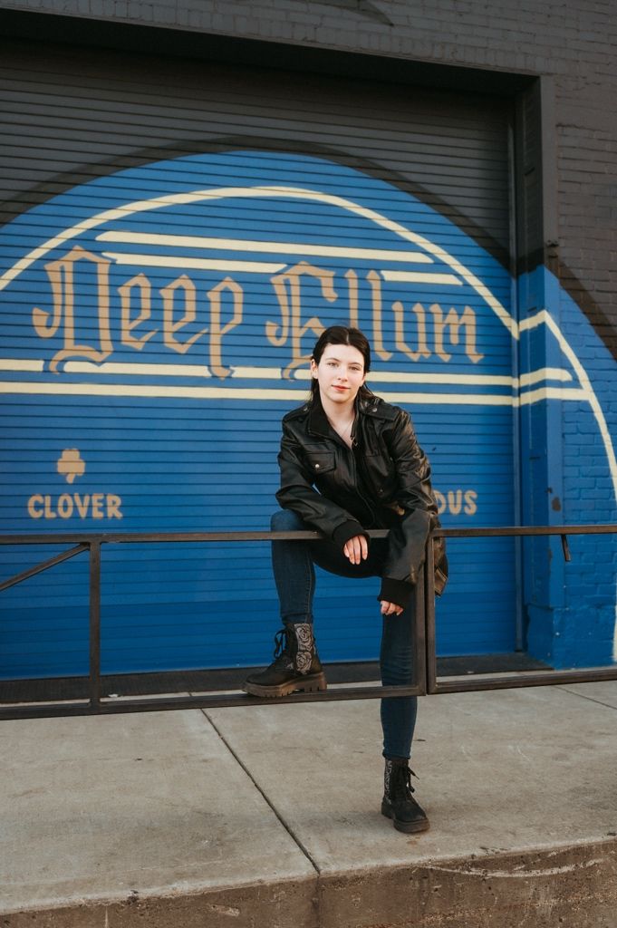 A person in black clothing poses against a blue garage door with vintage-style lettering and decorative design.