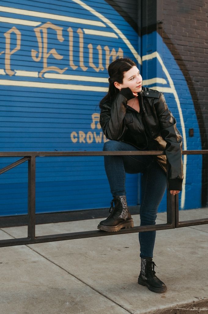 A person in black clothing crouches against a blue wall with text, creating a dynamic urban fashion pose.