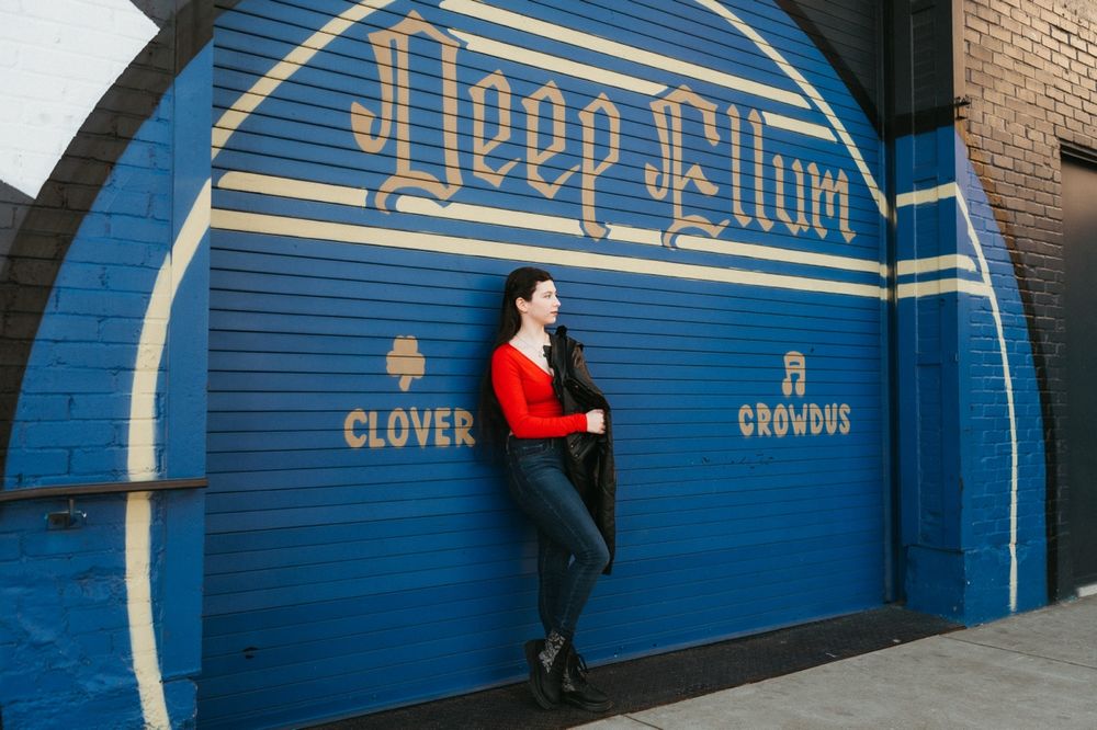 Someone poses against a bright blue garage door with 'Keep Willow' painted in stylized lettering at Clover Studios.